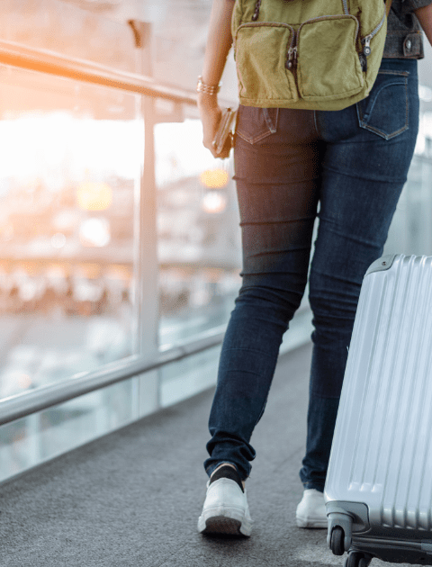 mulher de calça jeans e mochila verde com uma mala prateada no aeroporto